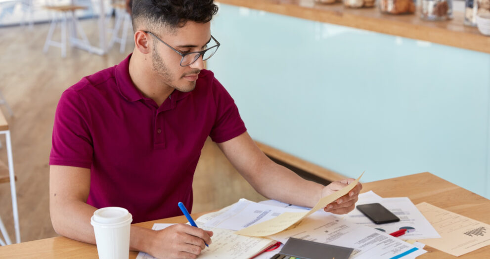 “A young man wearing glasses and a maroon polo shirt sits at a café table, reviewing documents and writing notes in a notebook. A coffee cup, smartphone, charts, and pastries are placed on the table around him.”