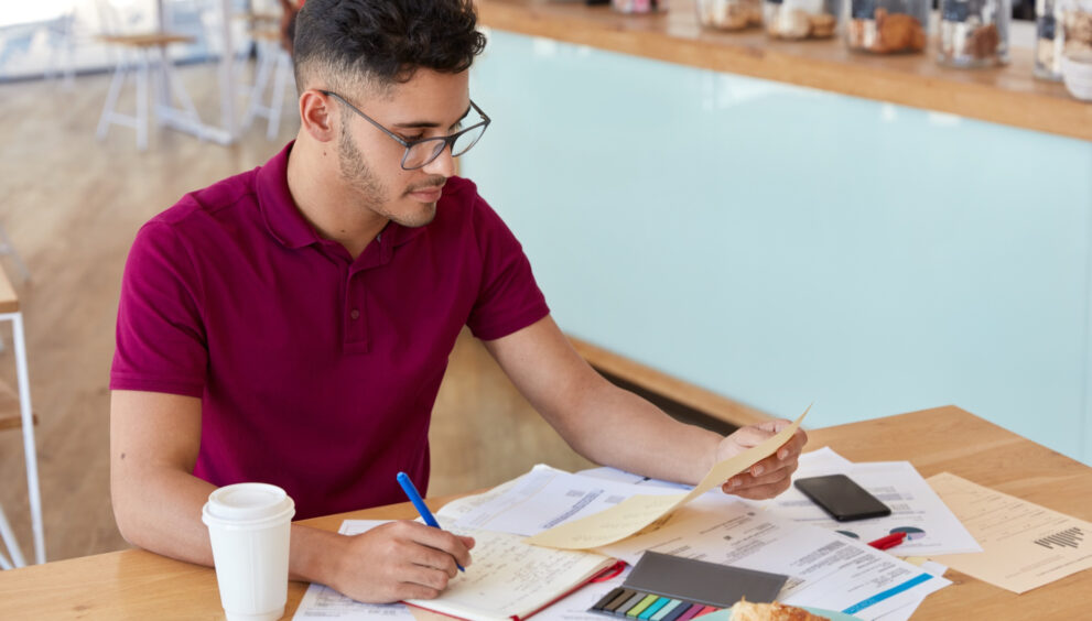 “A young man wearing glasses and a maroon polo shirt sits at a café table, reviewing documents and writing notes in a notebook. A coffee cup, smartphone, charts, and pastries are placed on the table around him.”